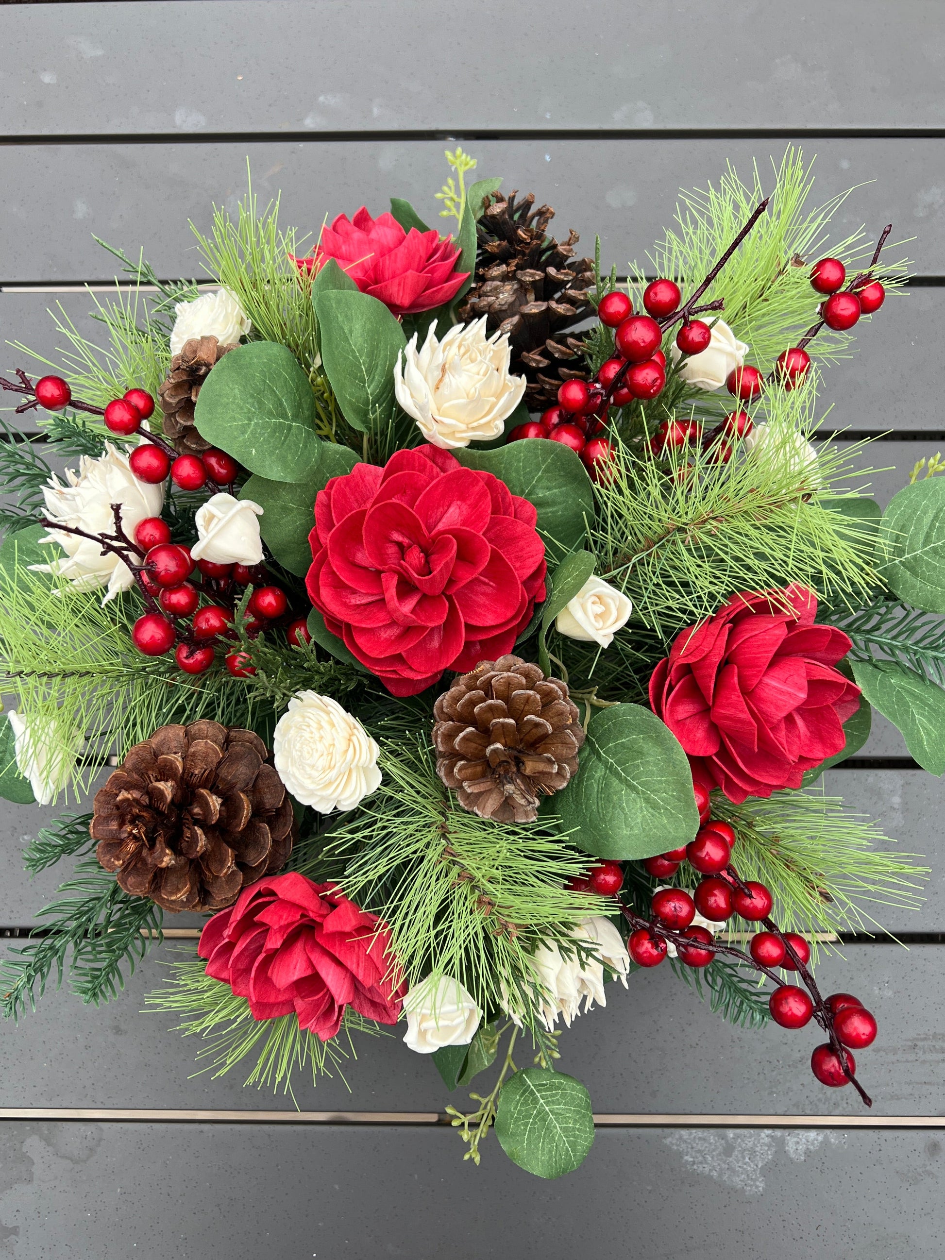 Christmas-themed floral arrangement with red flowers, greenery, and pinecones on a wooden surface.